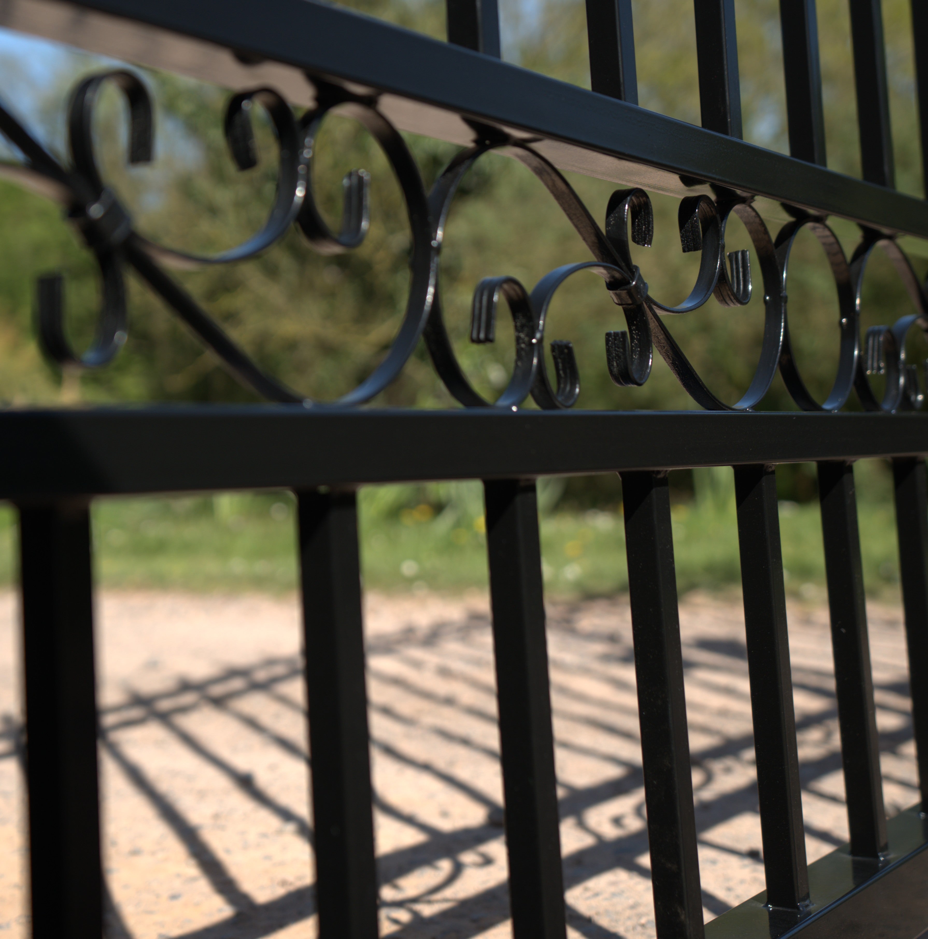 Close-up of a black metal gate with decorative patterns, blurred greenery in the background