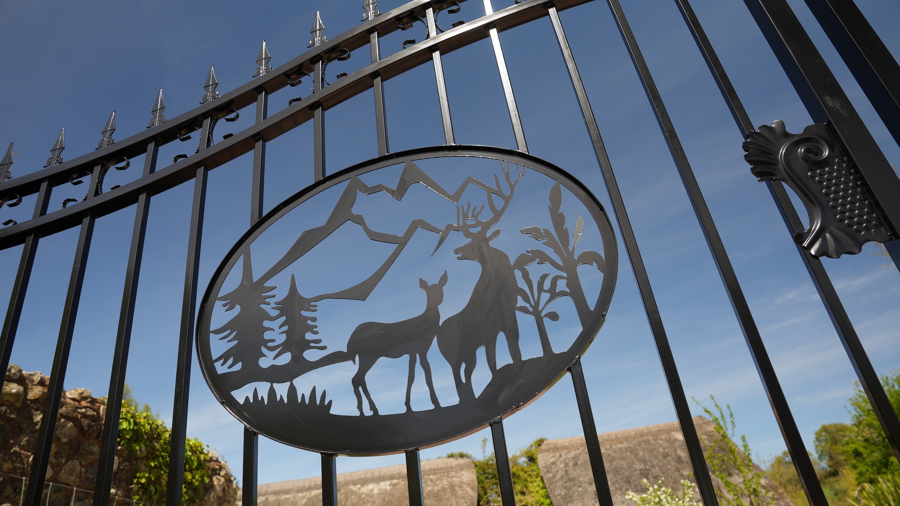 Decorative metal gate with deer and mountain design against a blue sky.